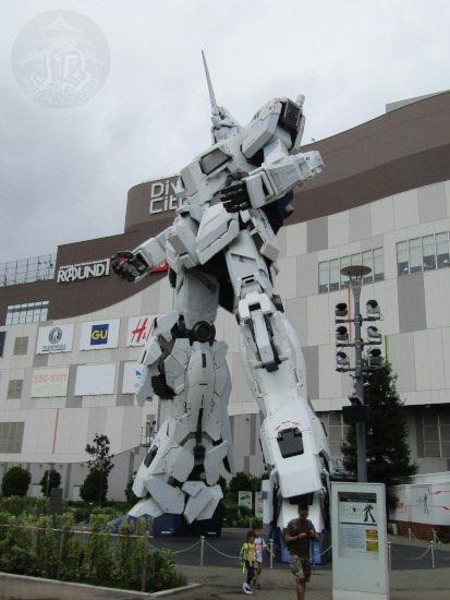 Gundan robot looking up to the sky. it is white and taller than the two-story shopping centre behind it.